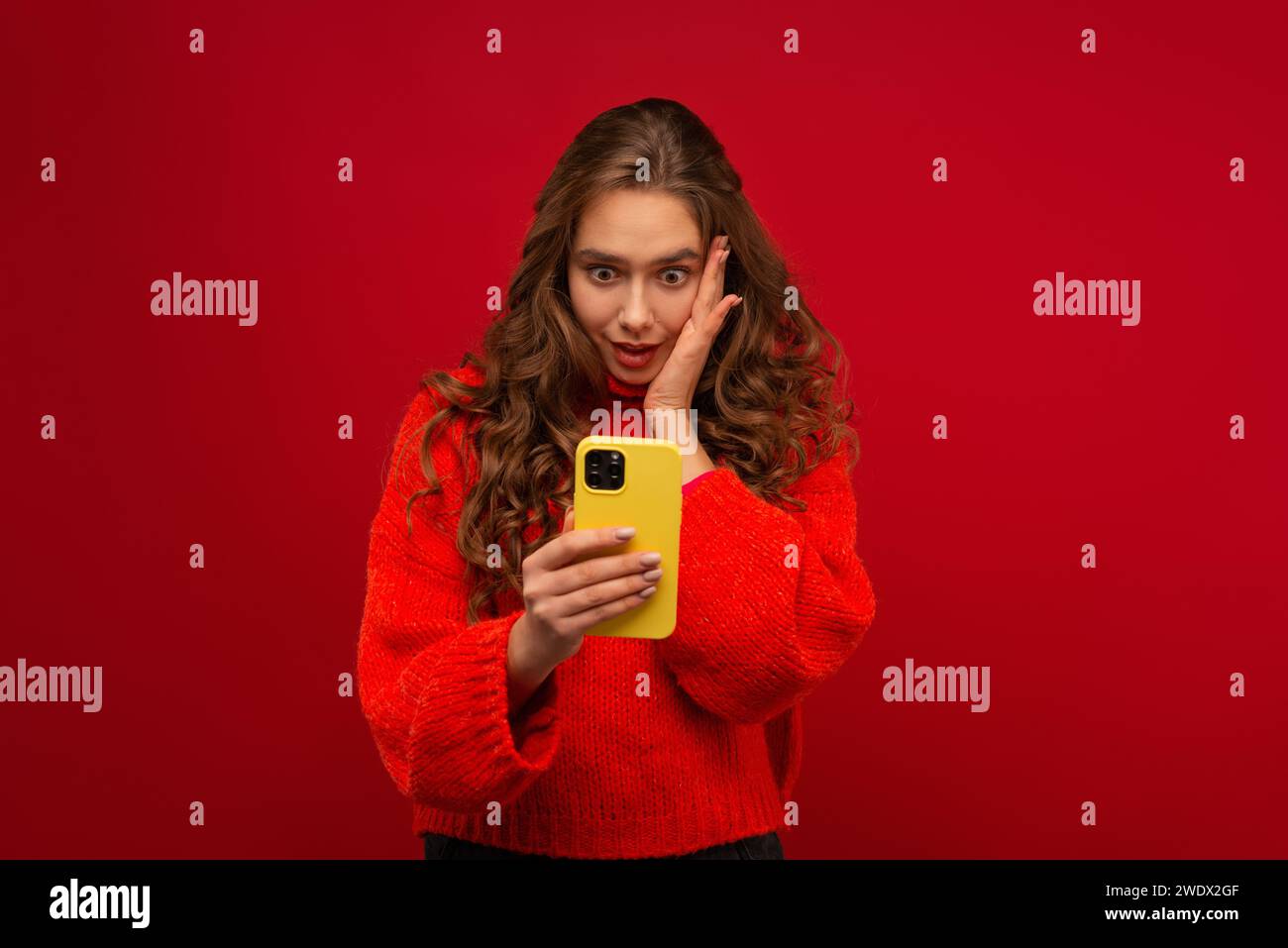 Portrait of a smiling emotional young woman in curly hair Generation Z in a red sweater using a modern smartphone in a studio shot red background Stock Photo