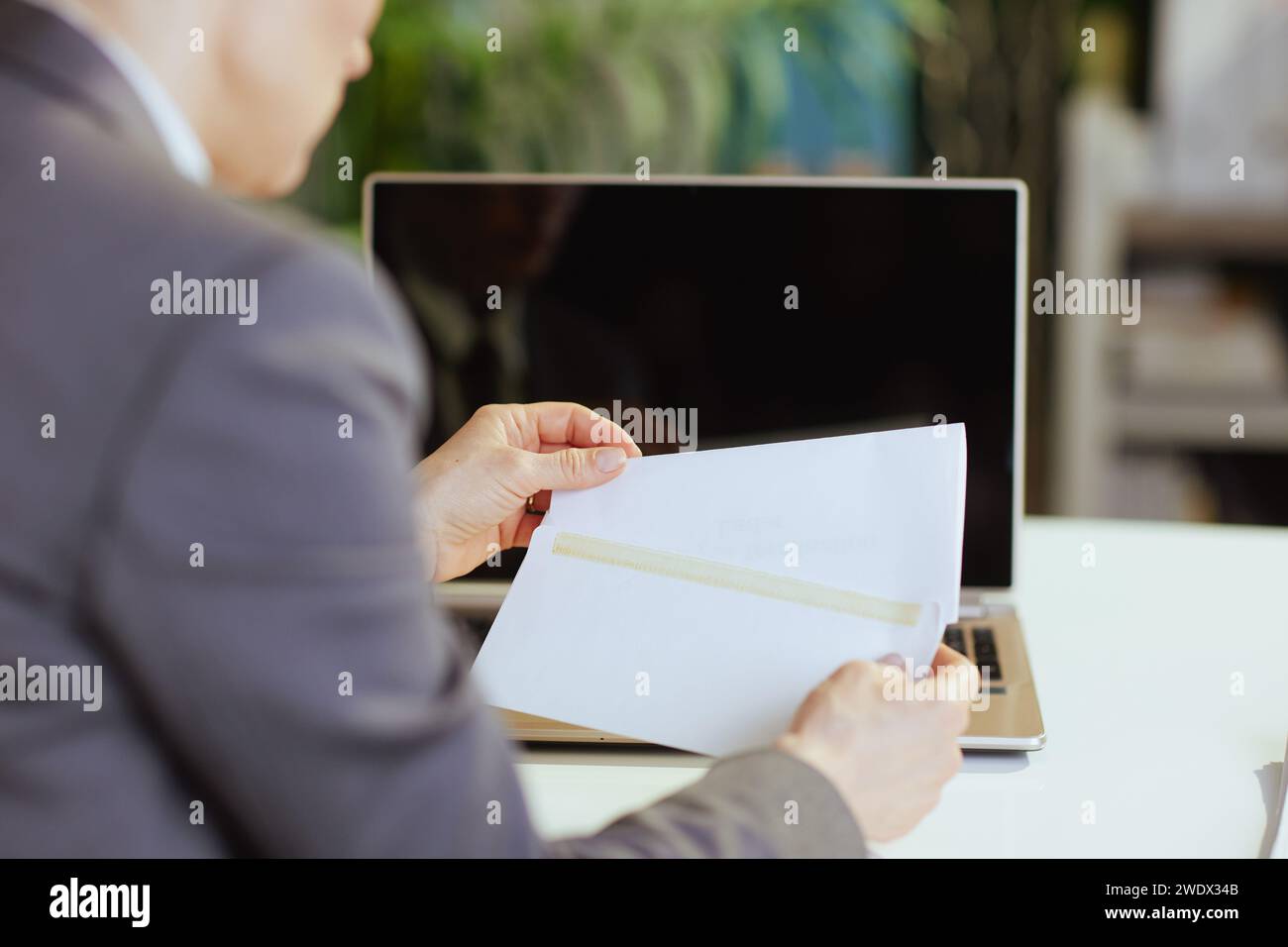 New job. Seen from behind modern middle aged woman worker in modern green office with laptop opening letter. Stock Photo