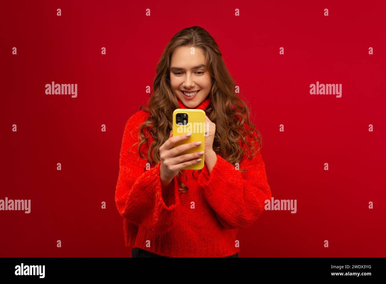 Portrait of a smiling emotional young woman in curly hair Generation Z in a red sweater using a modern smartphone in a studio shot red background Stock Photo