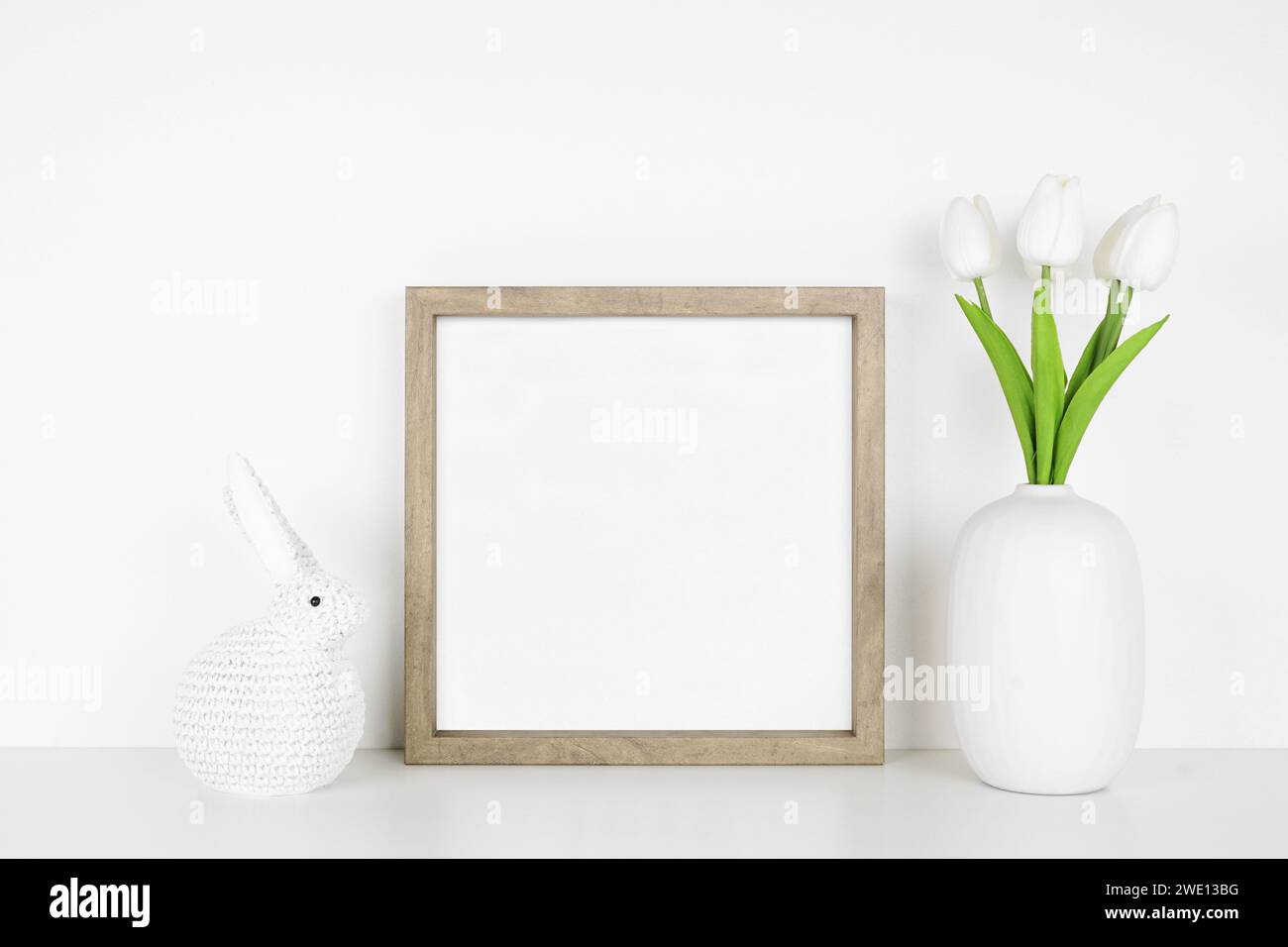 Mock up wood frame with Easter decor on a white shelf. Knit bunny and tulip flowers. Square frame against a white wall. Copy space. Stock Photo