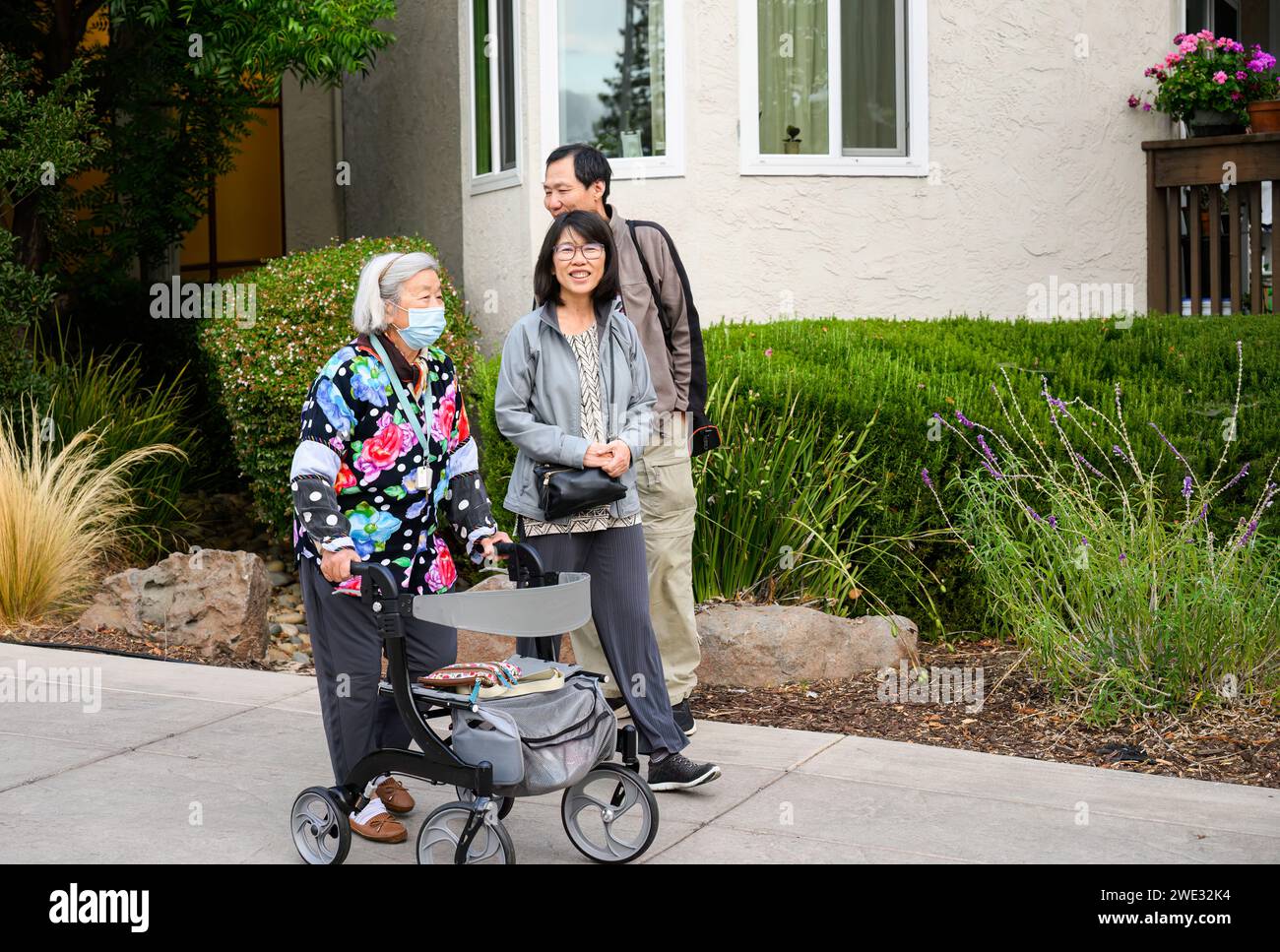 Senior woman walking with a mobility walker, with son and Daughter laughing and walking outside the retirement village. California. Stock Photo