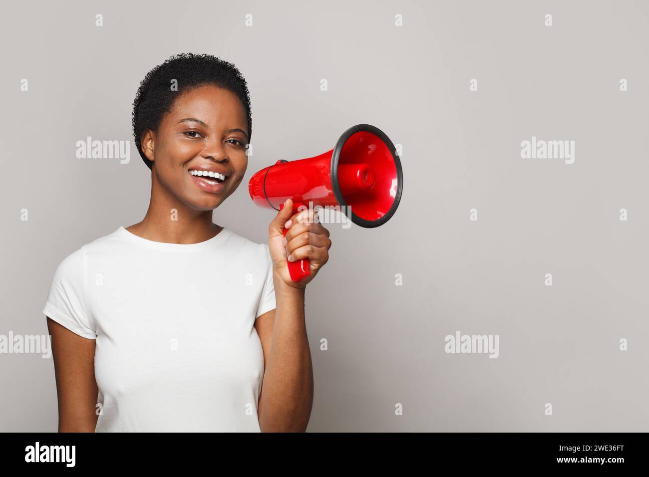 Joyful woman in white empty t-shirt holding loudspeaker against gray studio wall banner background Stock Photo