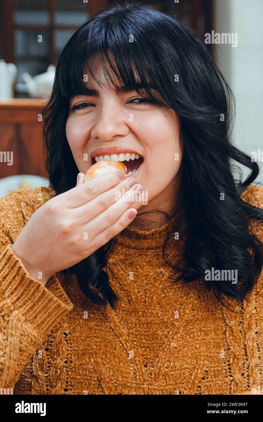 vertical image of young Latina with black hair and brown clothes, happy sitting in restaurant eating tequenos looking at camera, traditional Venezuela Stock Photo