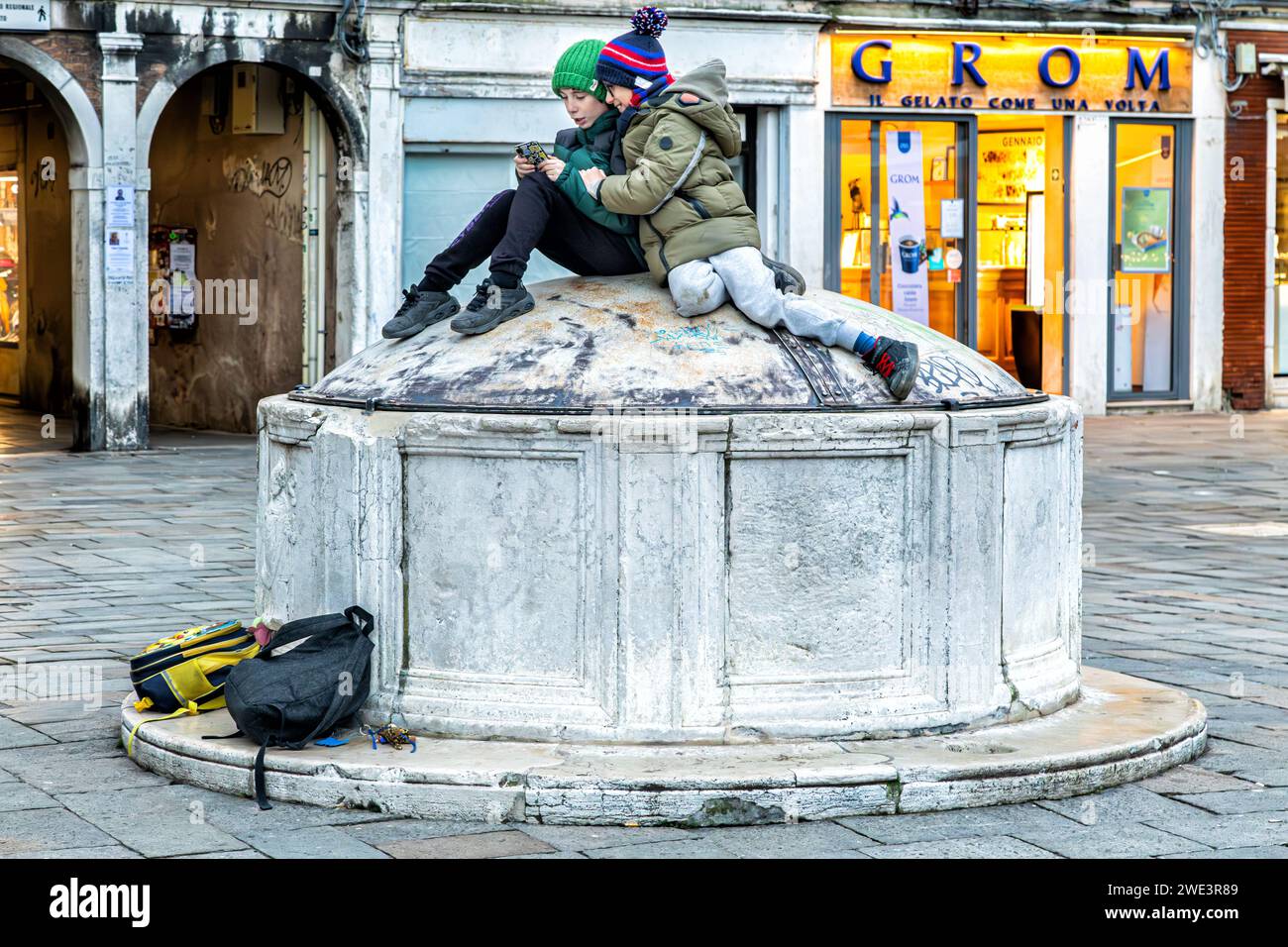 Kids playing with their phone while sitting atop a dwell in a campo (square) in Venice, Italy Stock Photo