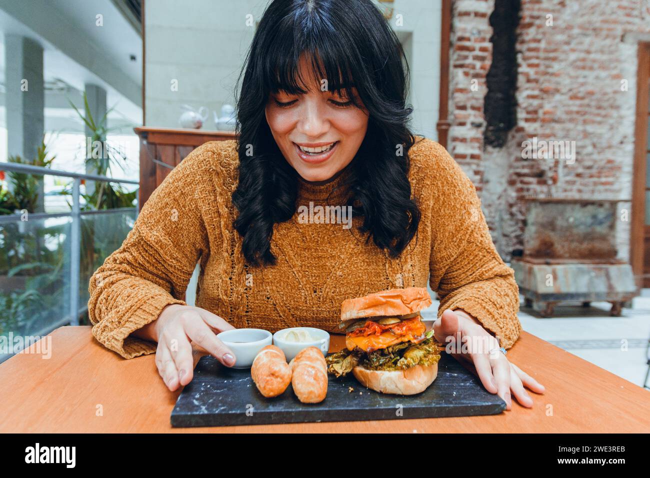front view of young hungry latin woman sitting in restaurant, happy with her lunch, going to eat tequeno and hamburger with salsa, she is smiling and Stock Photo