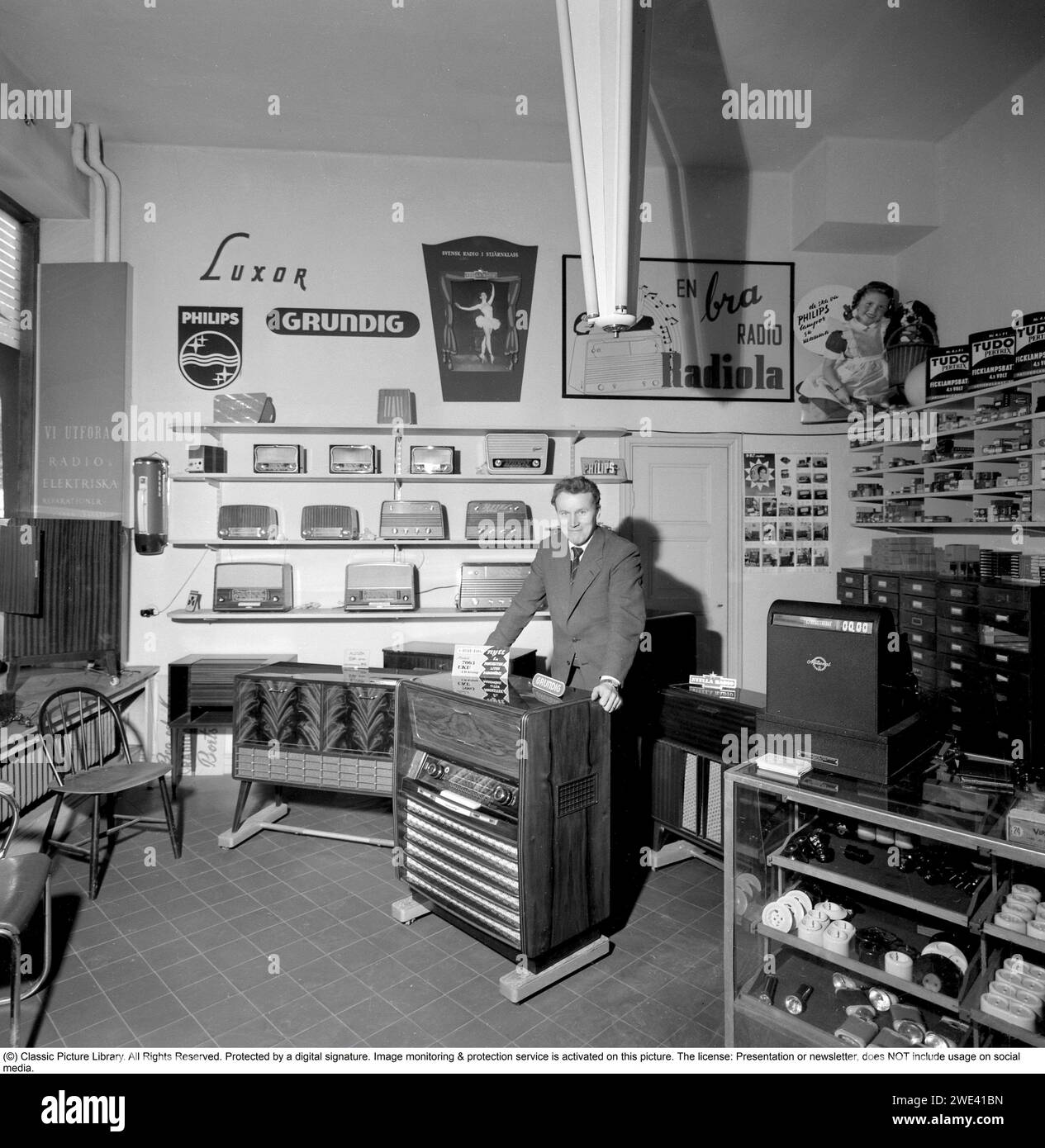 In the 1950s. Interior of a radio and tv store 1956. A salesman is seen at a Grundig radio gramophone Model 7063. On the shelfes are smaller transistor radios and advertisments for brands like Philips, Luxor and Radiola.   Conard ref 3210 Stock Photo