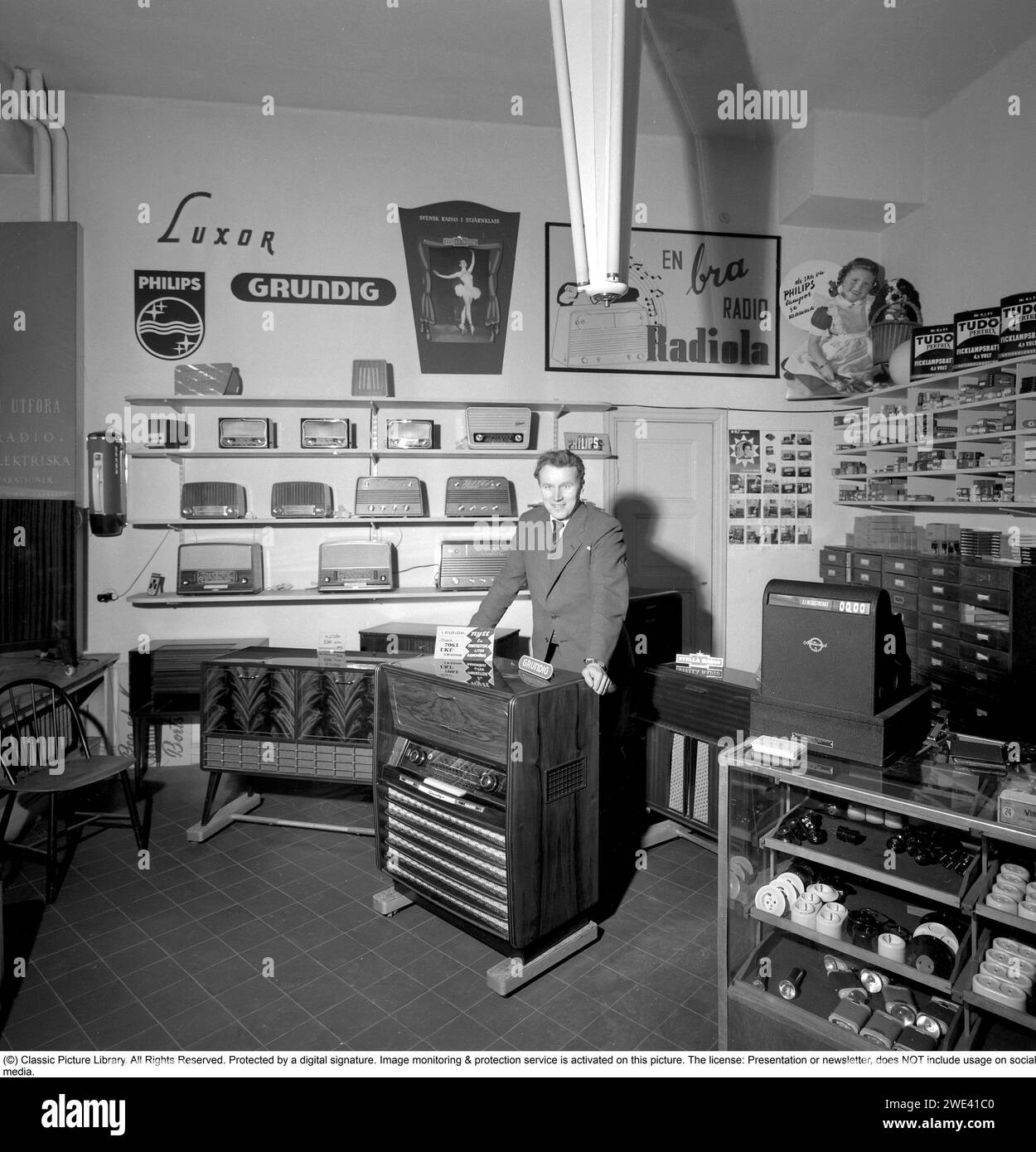 In the 1950s. Interior of a radio and tv store 1956. A salesman is seen at a Grundig radio gramophone Model 7063. On the shelfes are smaller transistor radios and advertisments for brands like Philips, Luxor and Radiola.   Conard ref 3210 Stock Photo