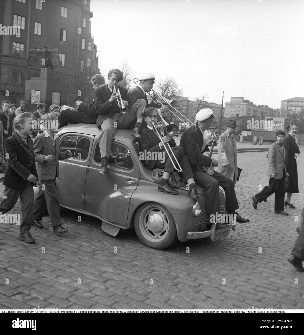 The student celebration in 1954. The students celebrate their completed matriculation and a band sits on top of a Renault 4CV which sags heavily under the weight of the orchestra. They pass the Aviator Monument at Karlaplan in Stockholm. Sweden 1954. Svahn ref SVA2 Stock Photo