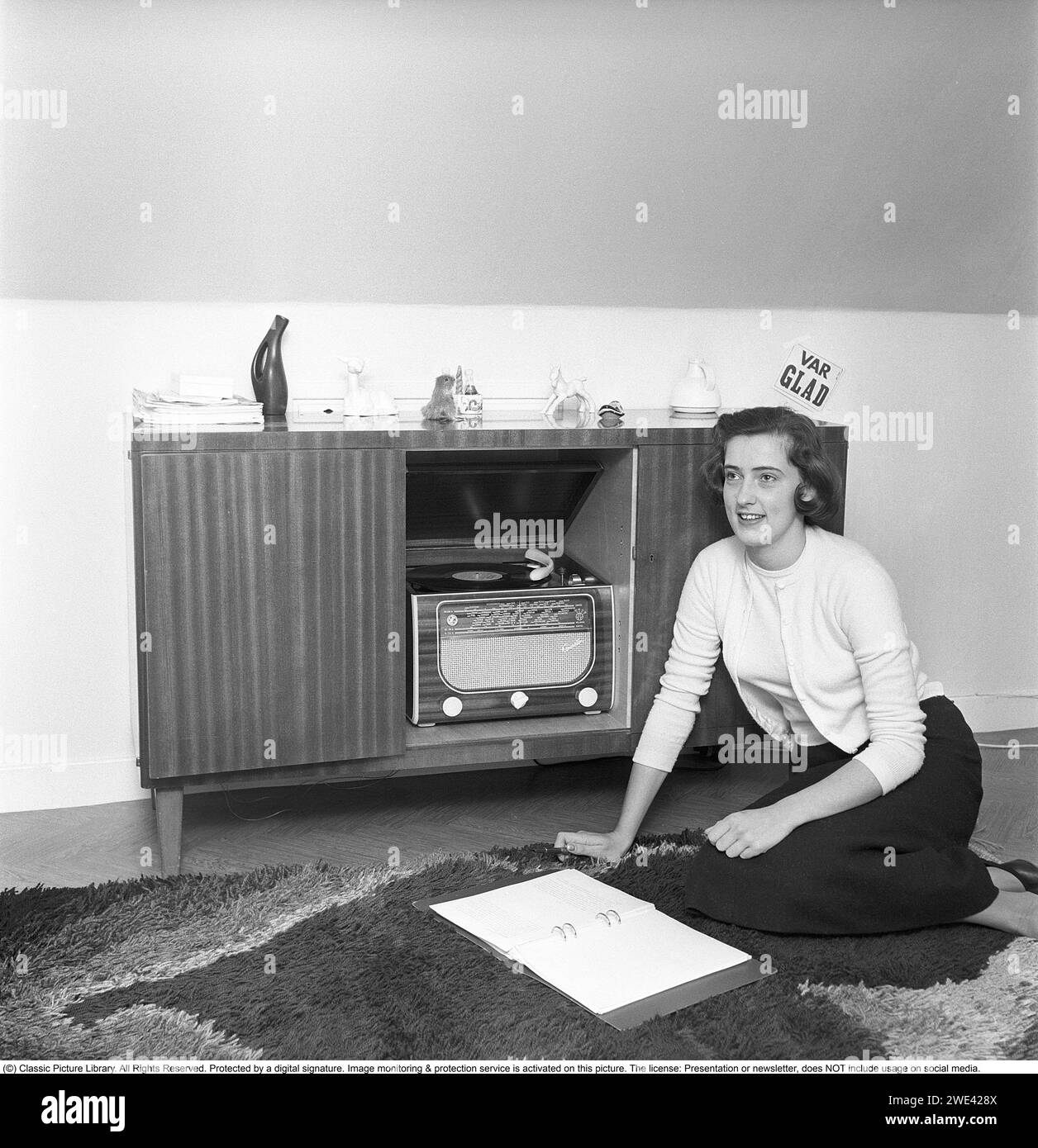 In the 1950s. A radio gramophone from the manufacturer Centrum radio. A combined piece of music furniture where a gramophone and radio were put together. The model became popular in the 1950s. The construction made them big and heavy, with more lavish details in the wooden construction. A young woman sits on the floor and listens to the music. Be happy reads the text on the sign on the wall. Sweden 1956. Svahn ref SVA1 Stock Photo
