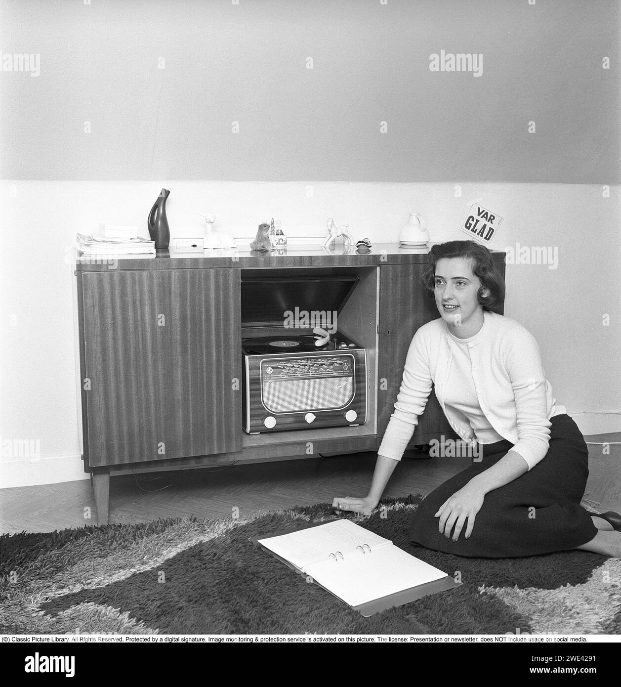 In the 1950s. A radio gramophone from the manufacturer Centrum radio. A combined piece of music furniture where a gramophone and radio were put together. The model became popular in the 1950s. The construction made them big and heavy, with more lavish details in the wooden construction. A young woman sits on the floor and listens to the music. Be happy reads the text on the sign on the wall. Sweden 1956. Svahn ref SVA1 Stock Photo