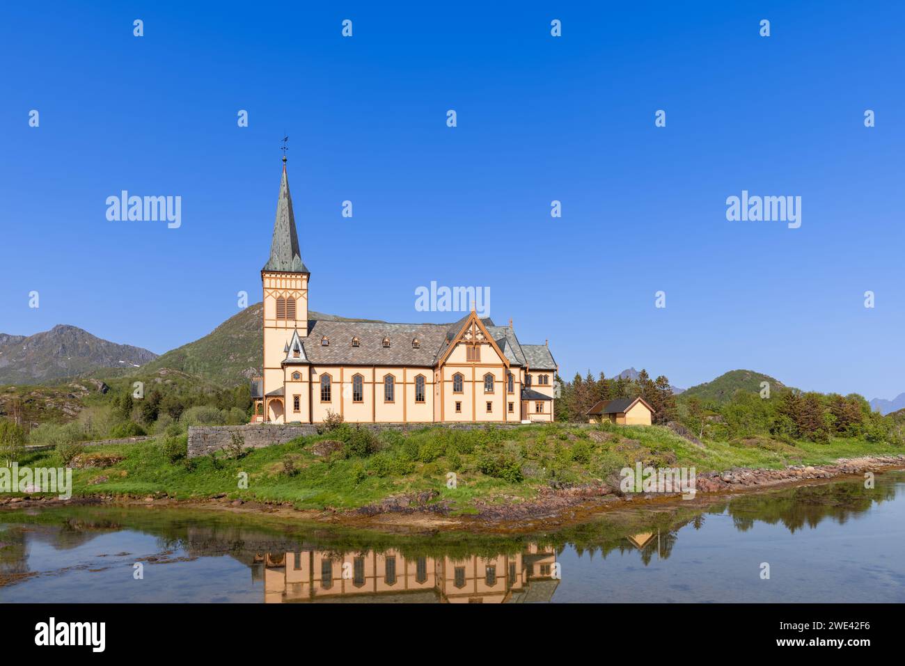 The Vagan Kirke in Lofoten, a stunning example of Gothic Revival architecture, reflects perfectly in the calm Norwegian waters under a vast blue sky Stock Photo