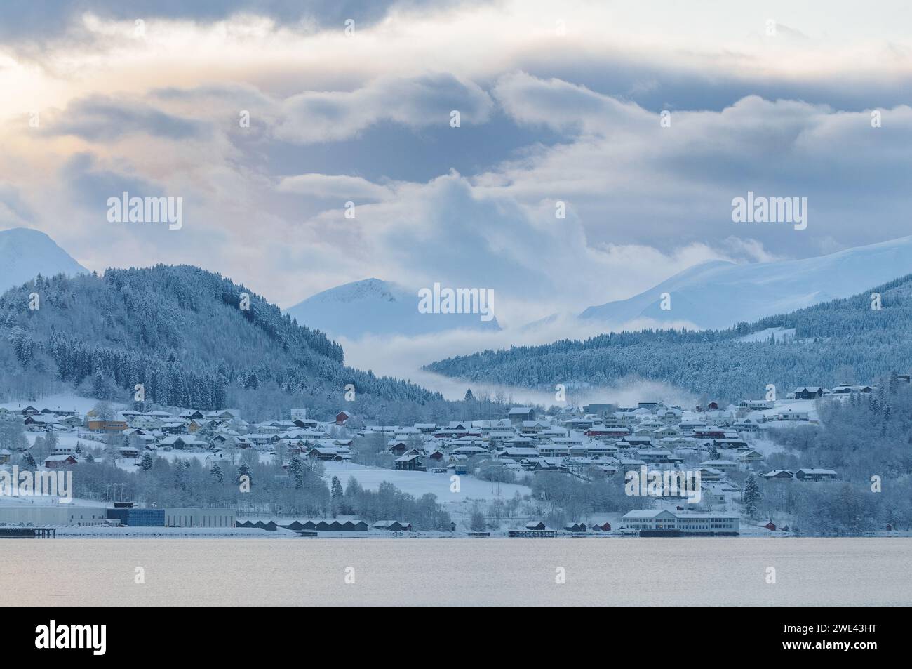 A serene snowy landscape showcases a quiet village beside the sea, enveloped by gentle mist and surrounded by forested mountains at dawn. Stock Photo