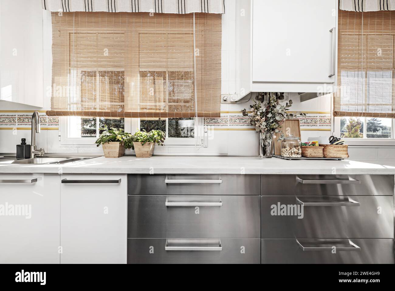 Front image of kitchen furnished with a combination of white base cabinets, stainless steel base drawers, white countertop and bamboo curtain on the w Stock Photo