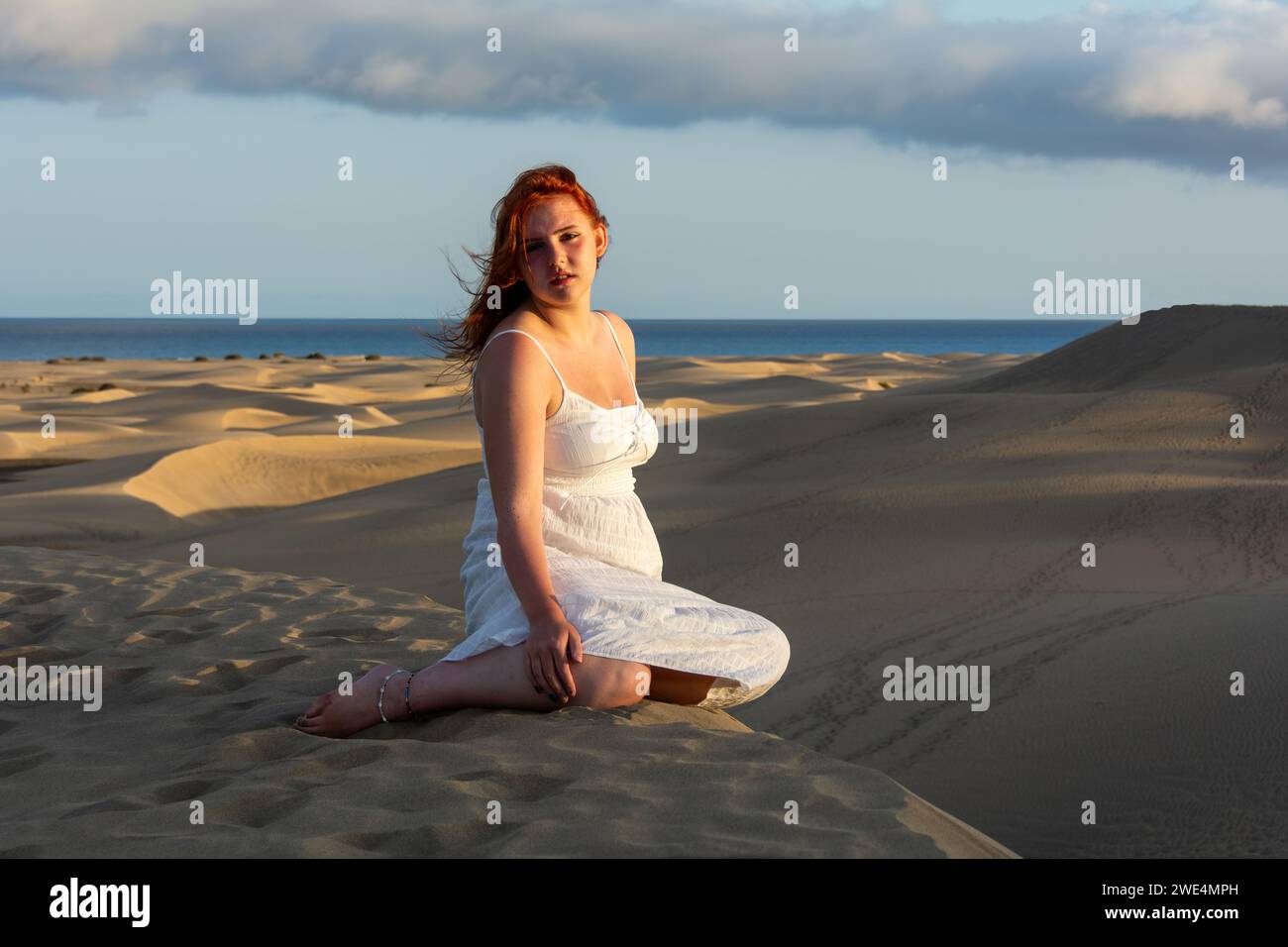 A young pretty red-haired girl in a white dress sits in the sand dunes in the late afternoon and enjoys the last rays of sunshine Stock Photo