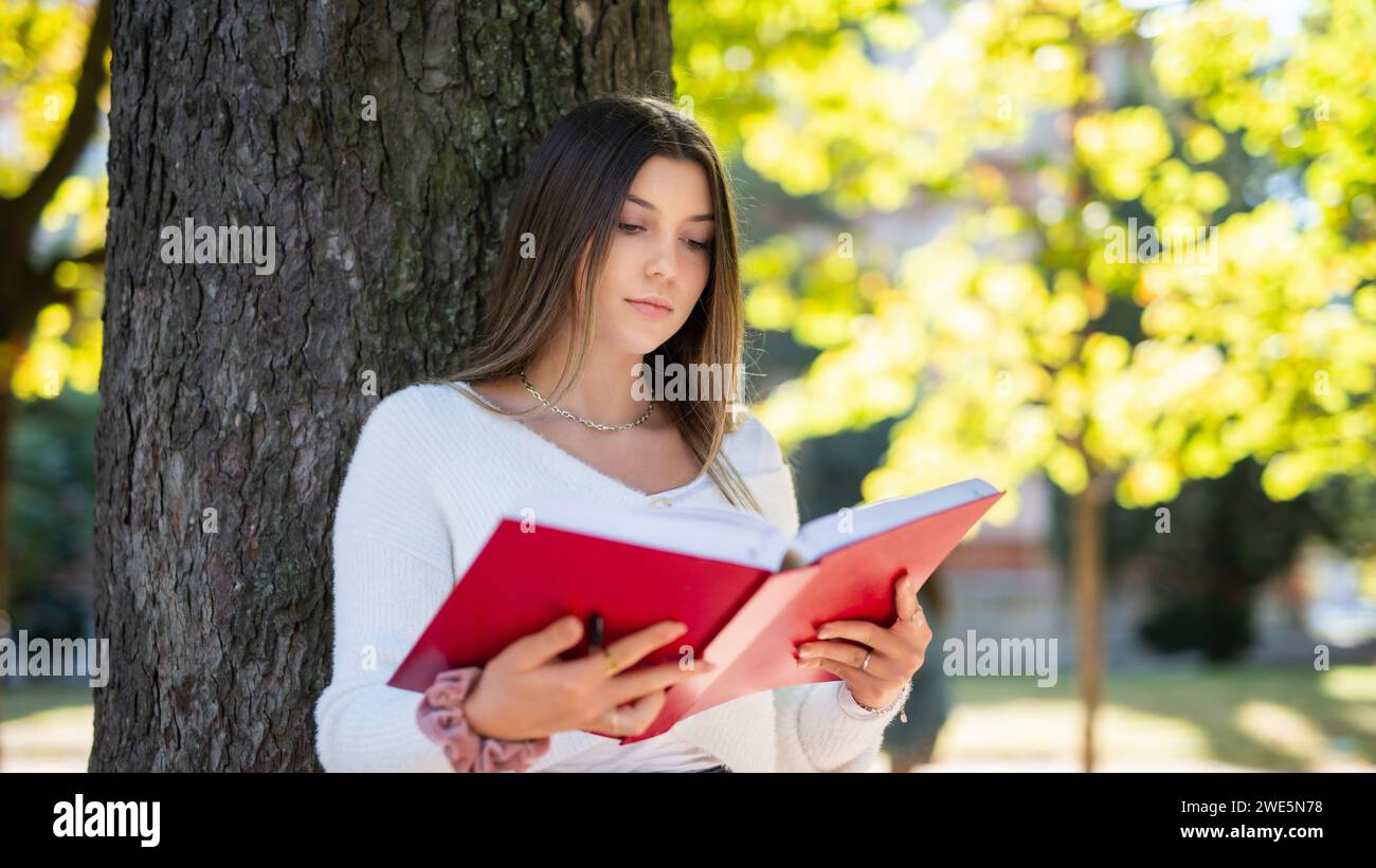 Woman reading a book while leaning on a tree in a park Stock Photo