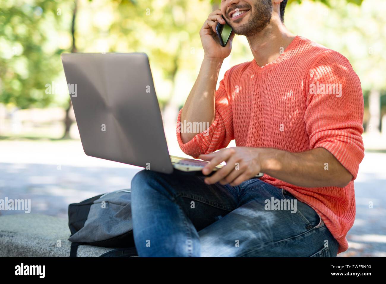 Young man using a laptop computer in a park while talking on his cellphone, young people and technology concept Stock Photo