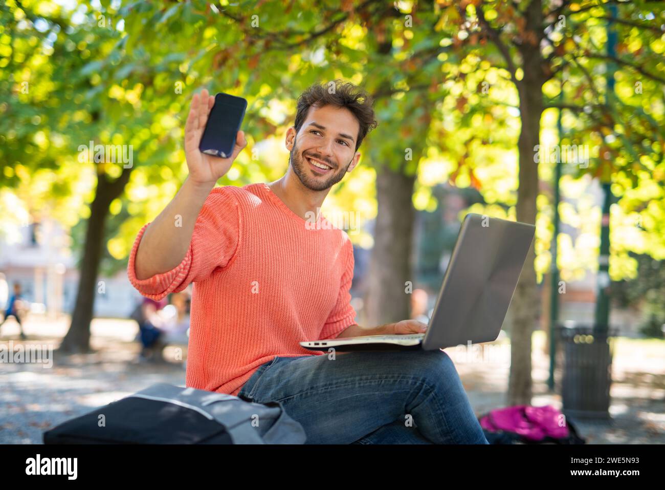 Young man using a laptop computer in a park while showing his cellphone, young people and technology concept Stock Photo