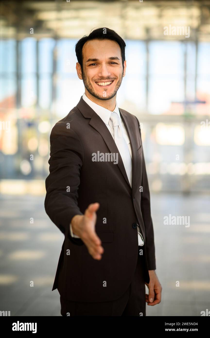 Professional young adult businessman in a stylish suit confidently offering a welcoming handshake in a modern office Stock Photo