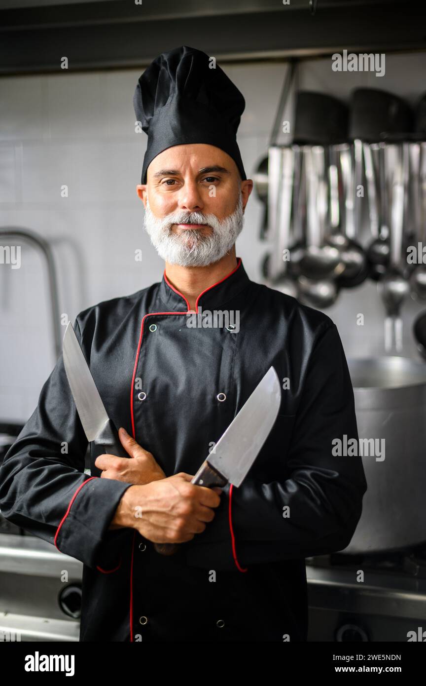 Professional male chef stands proud with crossed arms, holding two knives in a commercial kitchen Stock Photo