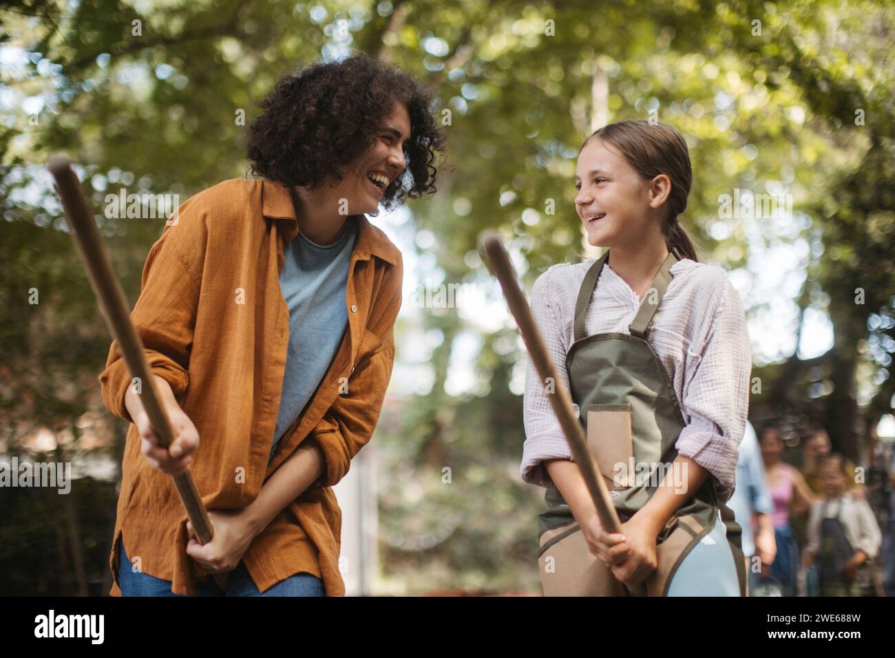 Woman and girl pretending to ride on broom sticks at community garden Stock Photo