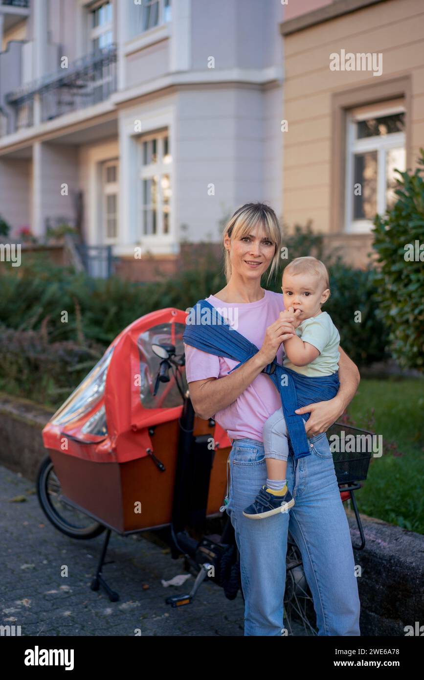 Woman carrying son in baby sling near cargo bike Stock Photo