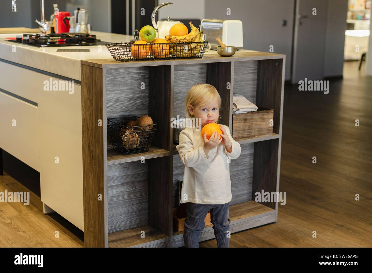 Girl holding orange fruit and standing near kitchen island at home Stock Photo