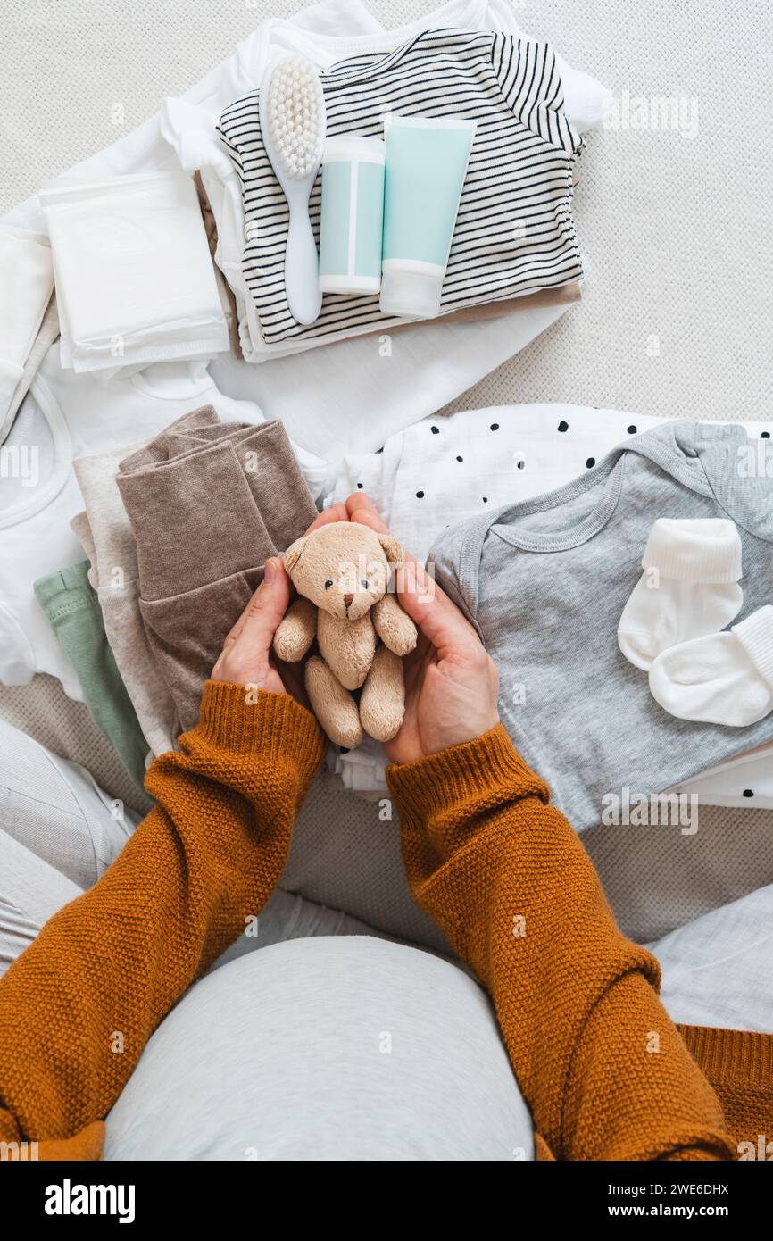 Pregnant woman holding stuffed teddy bear near clothes at home Stock Photo