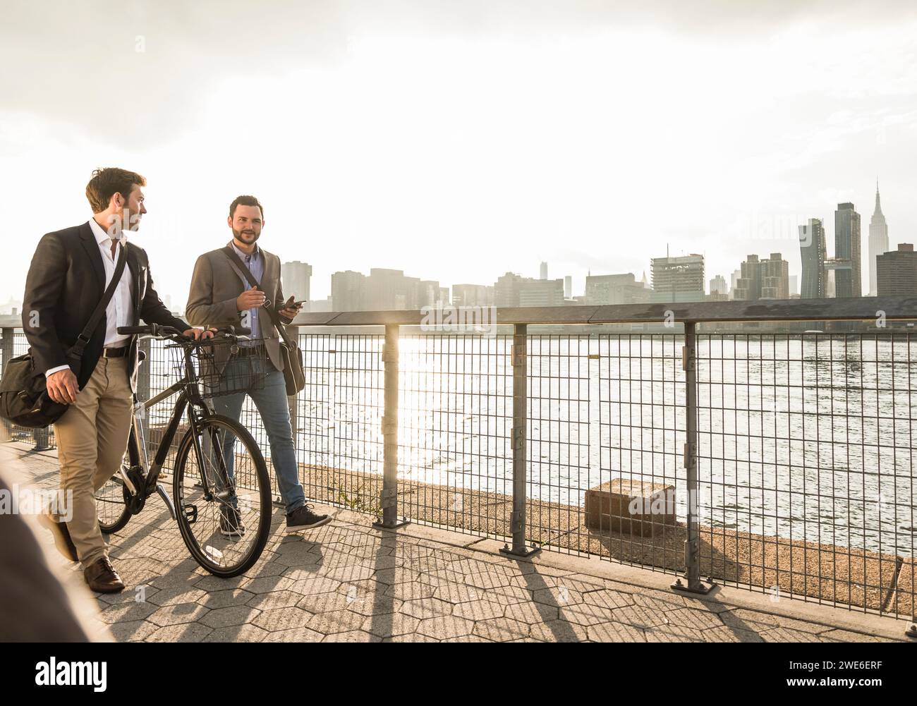 Businessman pushing bicycle and walking with colleague by river in New York City, USA Stock Photo