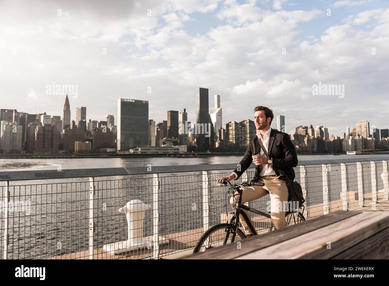 Businessman holding coffee cup and cycling by river in New York City, USA Stock Photo