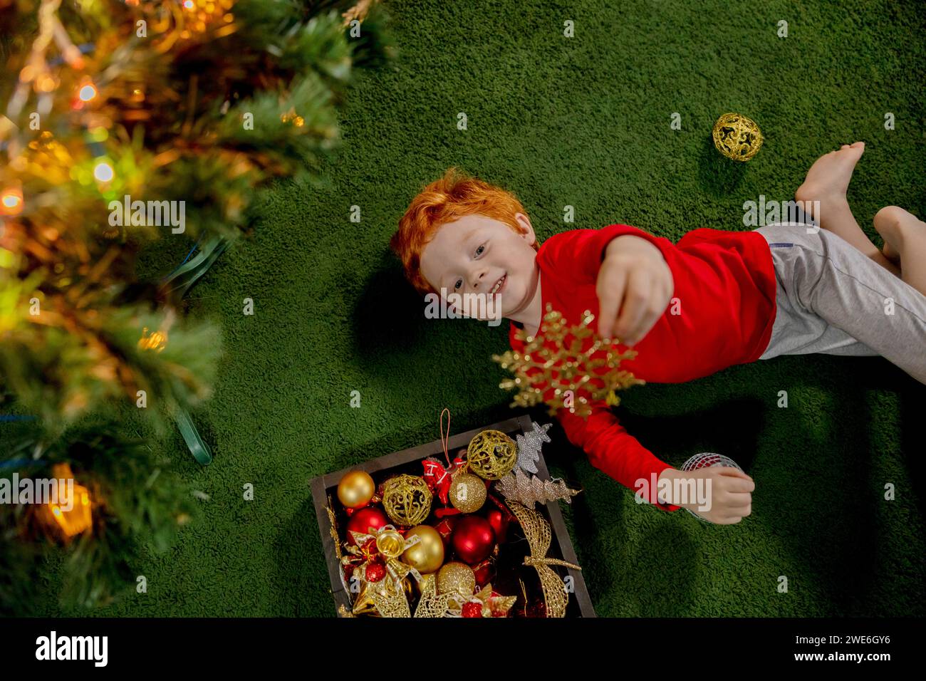 Happy redhead boy playing with Christmas ornament on green carpet Stock Photo