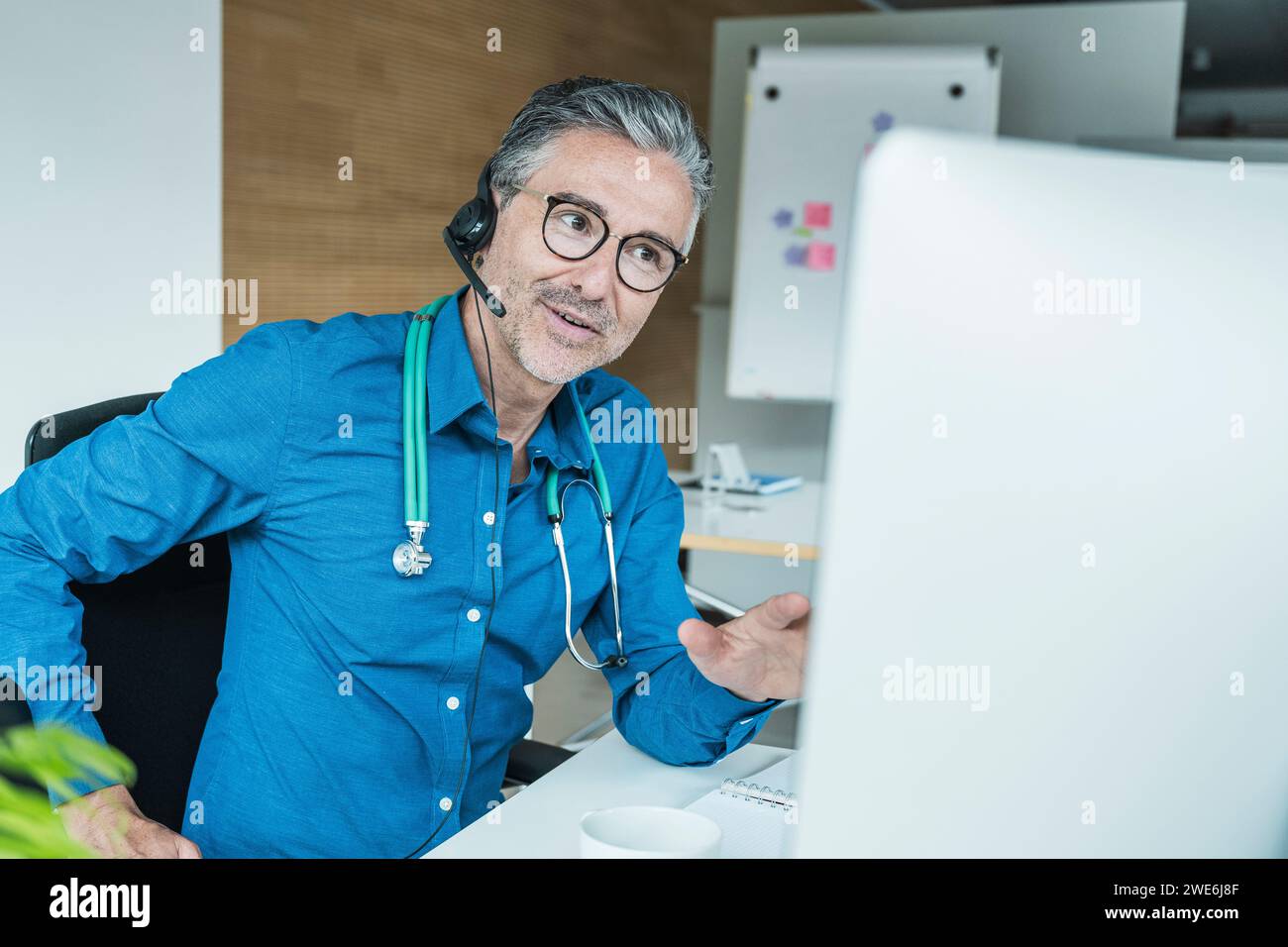 Mature doctor wearing headset and talking on video call through computer Stock Photo
