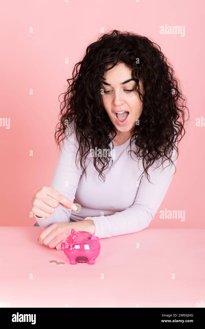 Surprised woman saving money in piggy bank against pink background Stock Photo