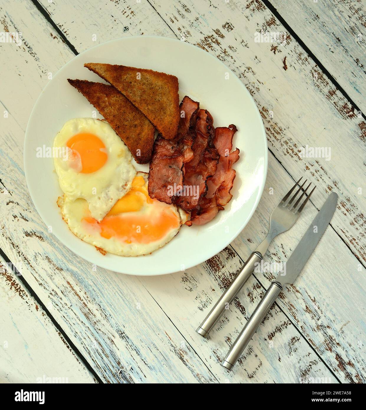 A plate of delicious breakfast, scrambled eggs with bacon, croutons and cutlery on a light wooden table. Top view, flat lay. Stock Photo