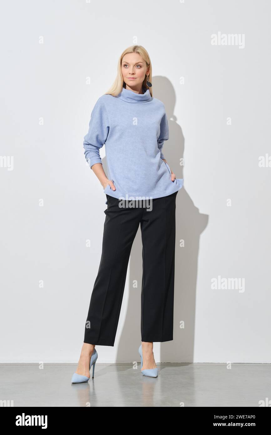 A modern fashion model posing with hands in pockets of classic black trousers wearing a stylish blue sweater and stilettos Stock Photo