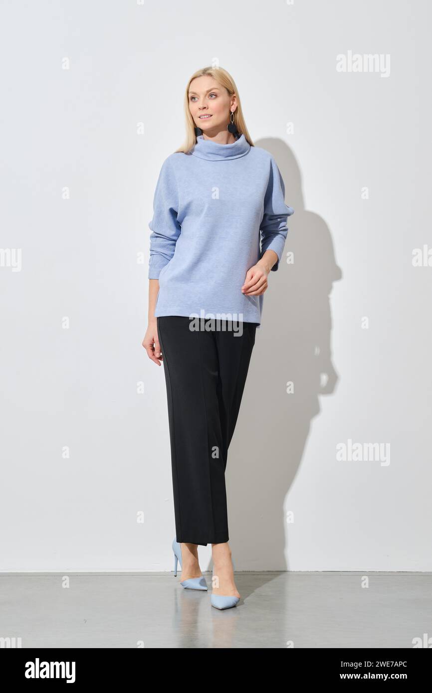 A modern fashion model poses in a relaxed stance, wearing a stylish blue sweater paired with classic black trousers, complemented by light blue heels Stock Photo