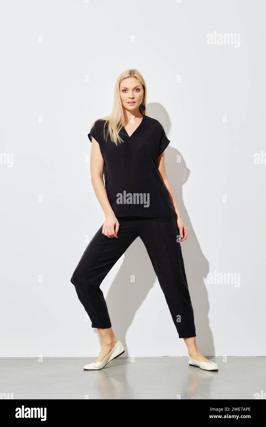 A fashion model wearing a black cotton outfit strikes a dynamic pose against a white wall, showcasing contemporary style and elegance Stock Photo