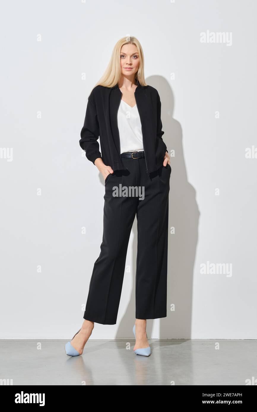 Confident woman in black pantsuit with hands in pockets posing next to white wall Stock Photo