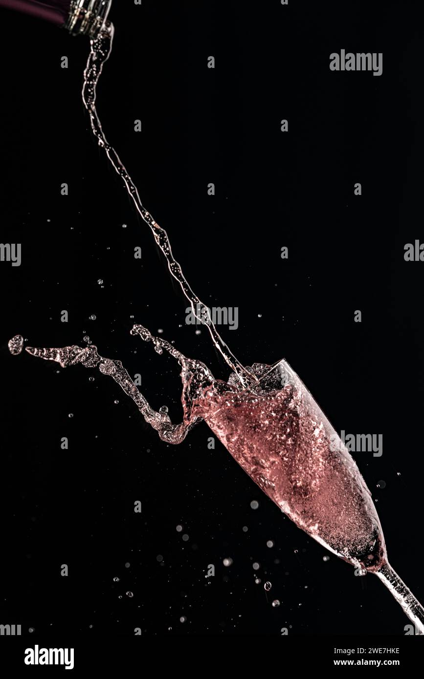 Rose champagne pours from a bottle into a champagne glass, drops and splashes fly, black background Stock Photo