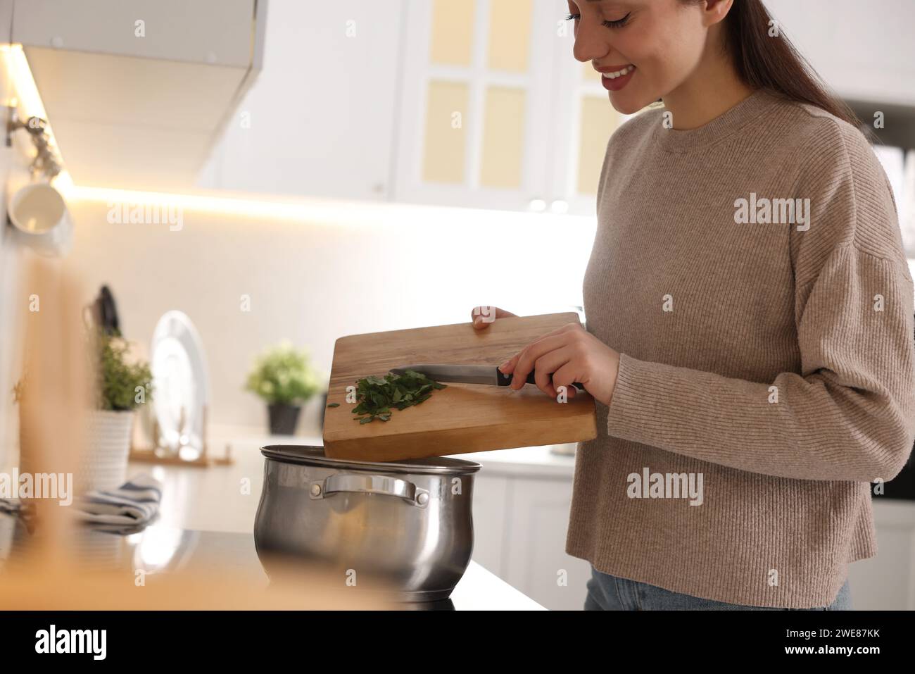 Smiling woman adding cut parsley into pot with soup in kitchen, closeup Stock Photo