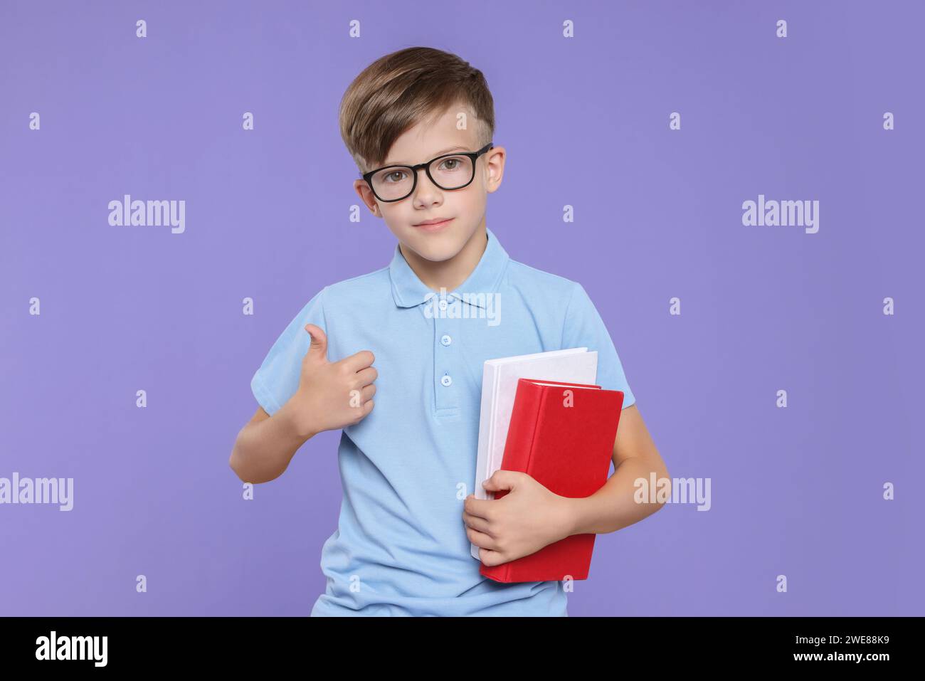 Cute schoolboy in glasses holding books and showing thumbs up on violet ...
