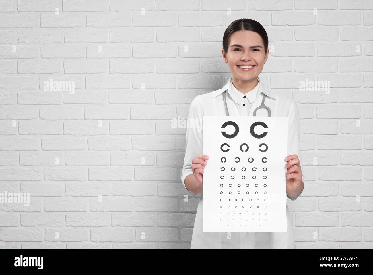 Ophthalmologist with vision test chart near white brick wall, space for text Stock Photo