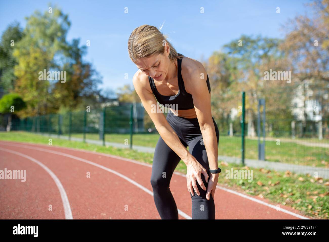 Fit woman in athletic wear rests with hands on knees after running on
