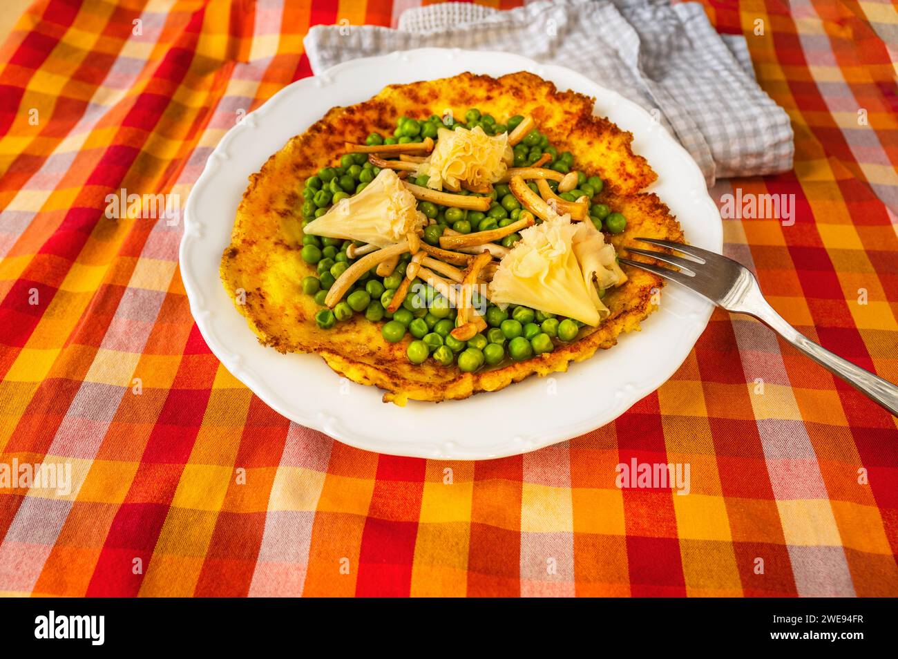Egg omelette with boiled pea, fried shimeji mushroom and rose-shaped swiss cheese on white plate with fork on red-orange checkered tablecloth, towel, Stock Photo