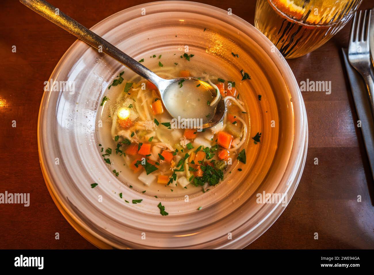 Portion of beef broth with sliced vegetable and noodle in plate, spoon, drink on table, closeup. Stock Photo