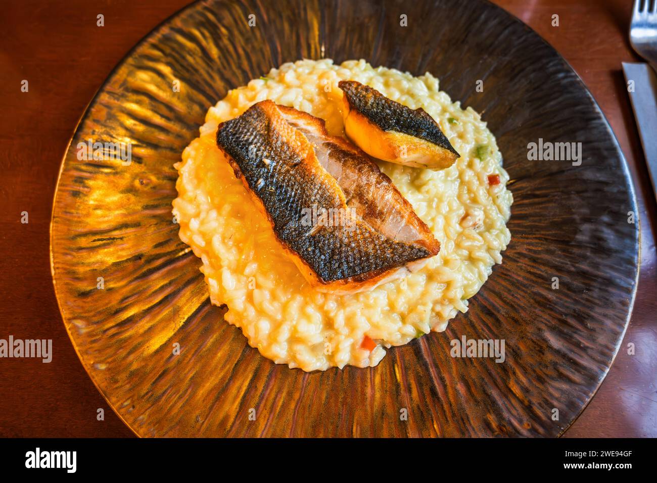 Roasted fish fillet (sea bass) on risotto with vegetable on decorative plate on restaurant table, closeup. Stock Photo