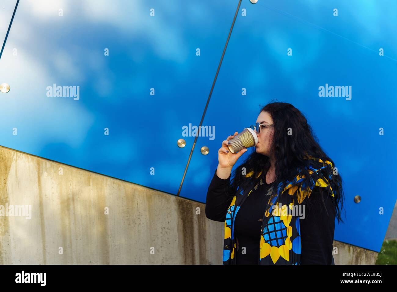 Beautiful young brunette woman with long curly hair, dressed in a black, blue and yellow waistcoat, drinks coffee from a paper cup on a background of Stock Photo