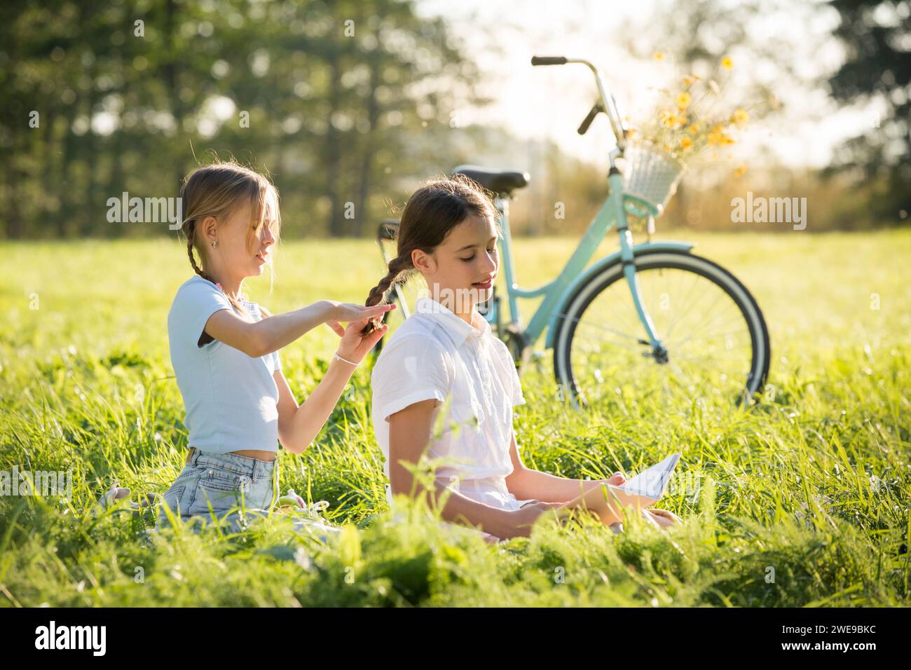 Two teenage girls spend time on green grass lawn in park, braid pigtails and tails for each other, read books, enjoy summer and vacations Stock Photo