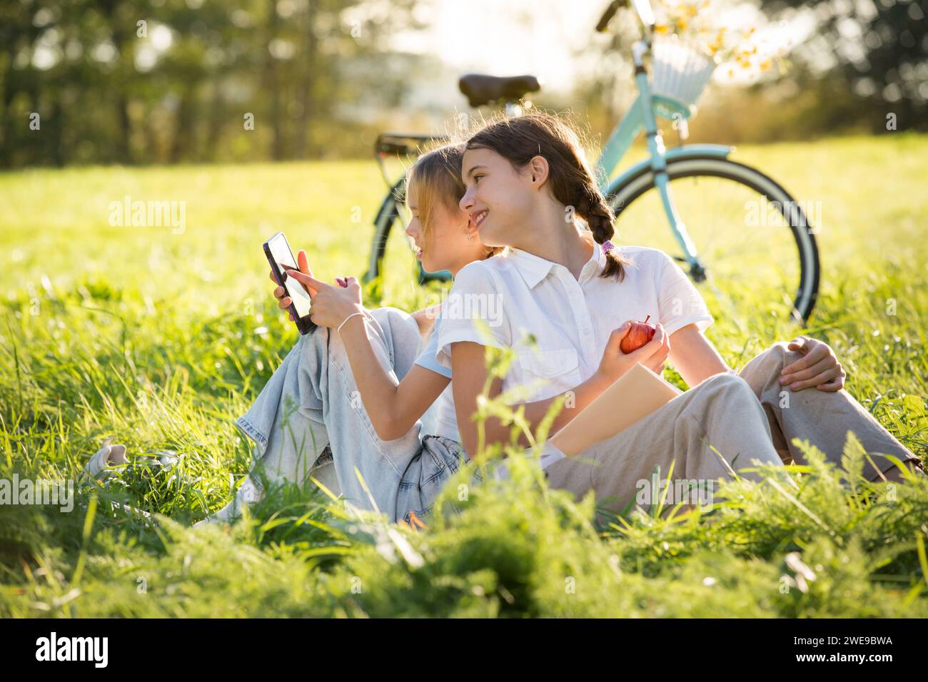 Two girls spend time on green grass lawn in park together, scroll through smartphones, read books, eat apples, enjoy summer and vacations Stock Photo
