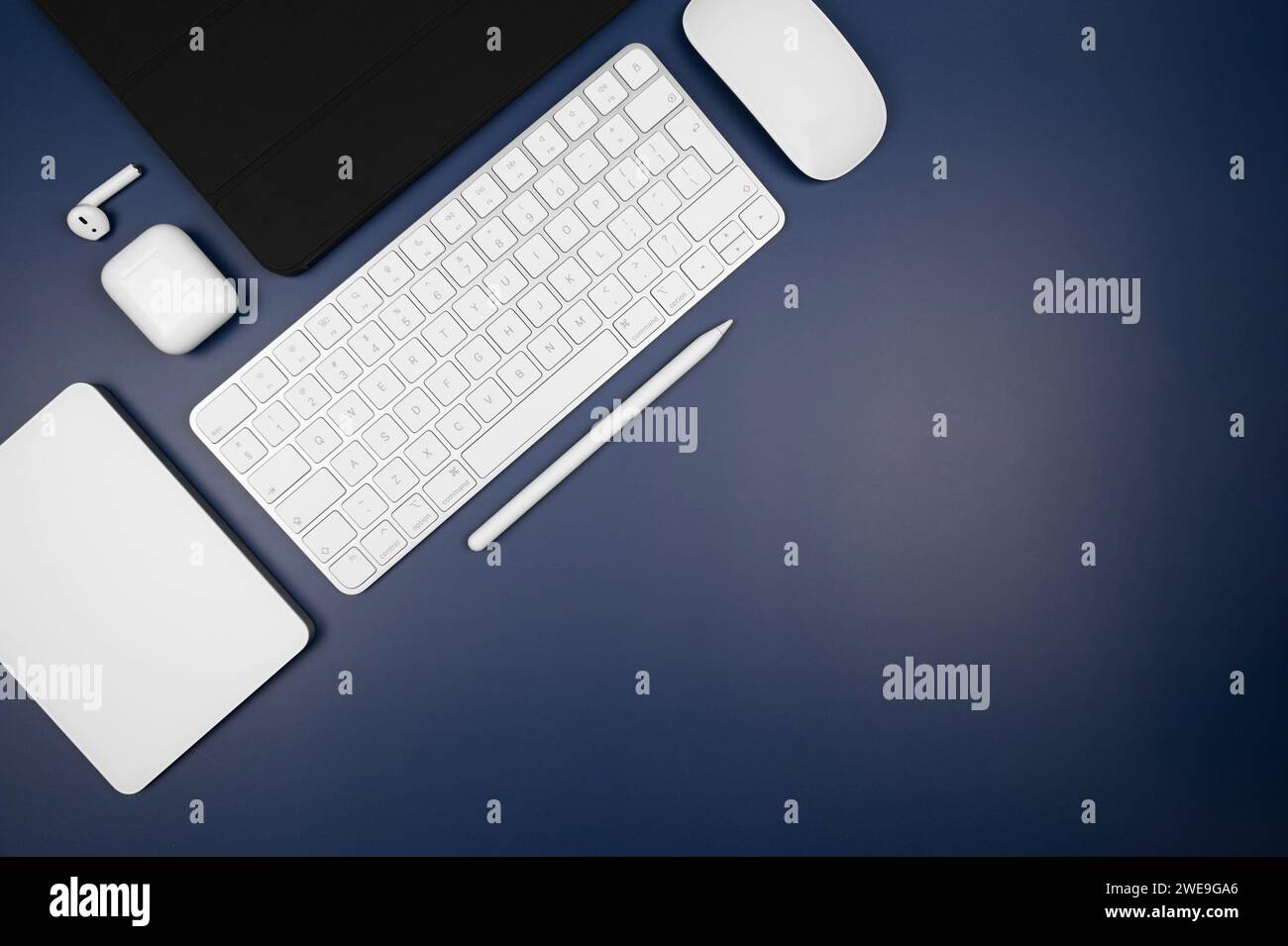 Top view of white keyboard, touchpad, mouse, earphones case and pen on dark blue background. Modern office flat lay, black tablet case, copy space. Stock Photo
