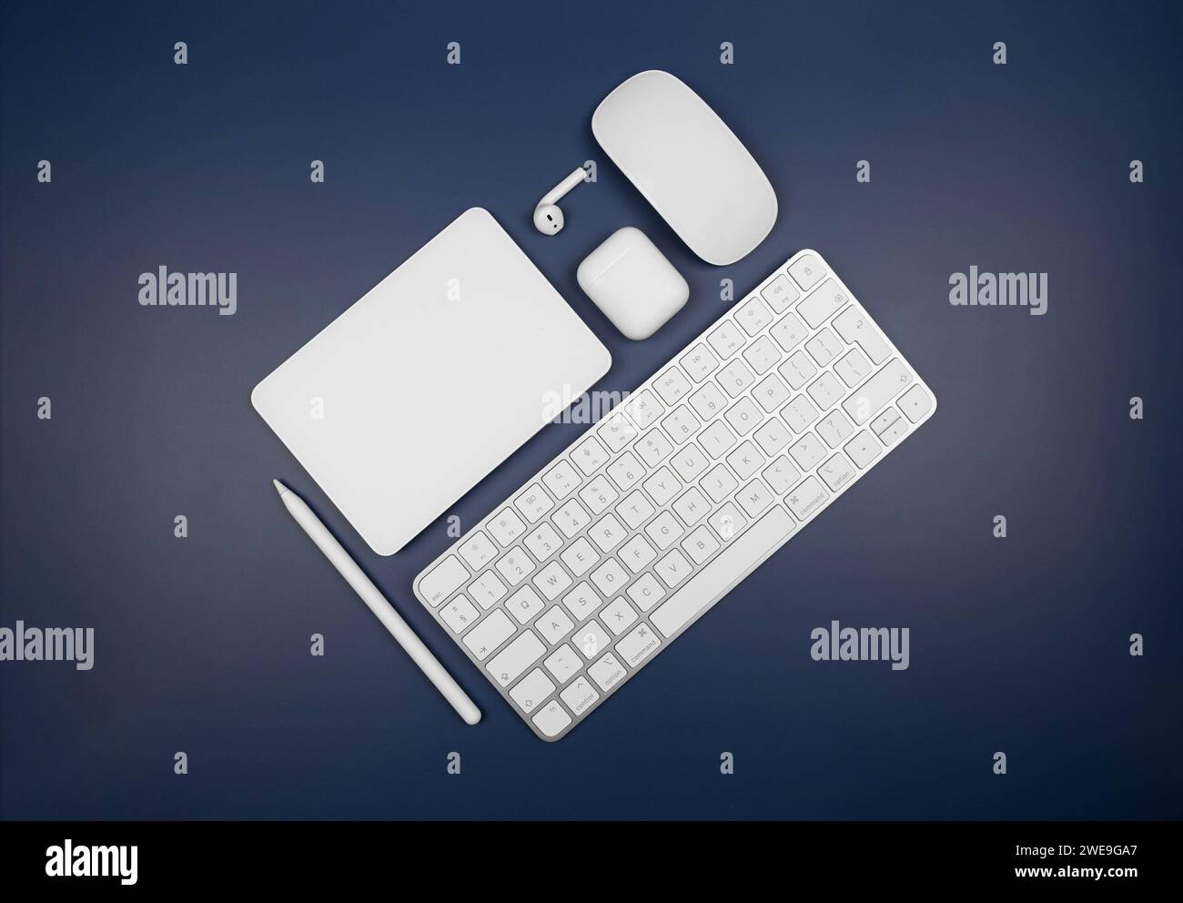 Top view of white keyboard, touchpad, mouse, earphones case and pen on dark blue background. Modern office flat lay, copy space. Stock Photo